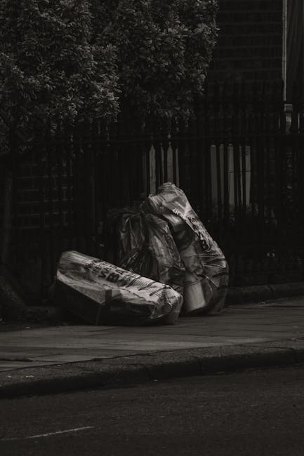 On a city sidewalk, three large black garbage bags filled with waste rest against a wooden fence and near a metal railing, with trees and foliage visible in the background. The bags appear to be made 